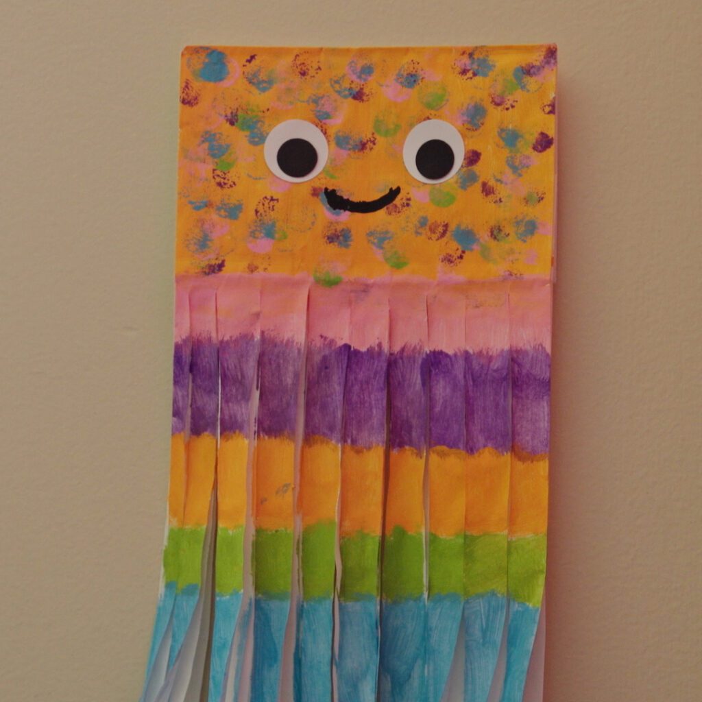 A photograph of a paper bag that a child colorfully decorated into a jelly fish. It's face has googly eyes and a smile. 