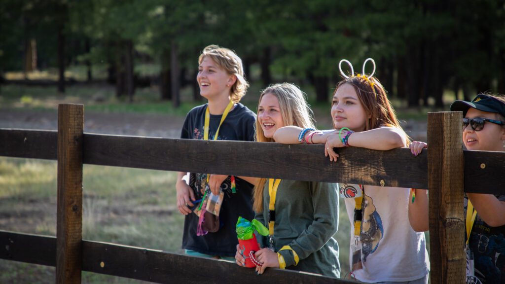 Four campers lean over a fence at Camp Rainbow. They are all smiling at something in front of them.