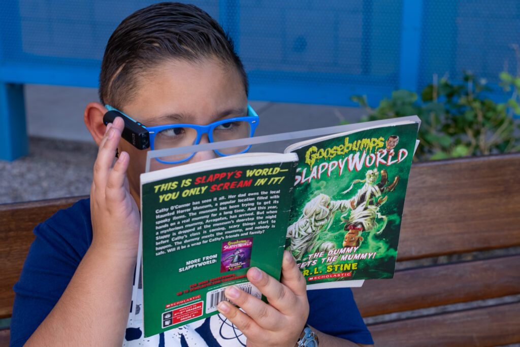 Christian reads his Goosebumps book with his OrCam MyEye2 glasses. He touches the side of the device which rests on the frame of his glasses. 