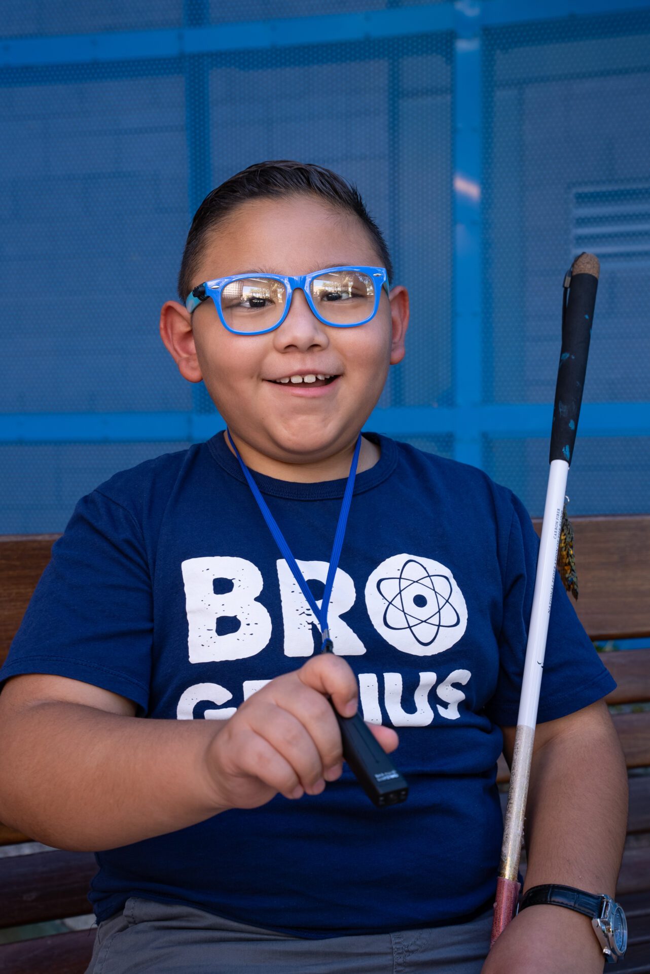 Christian poses for a photo. He wears blue glasses. He holds his OrCam MyEye 2 device in his hand. His shirt reads "Bro Genius"