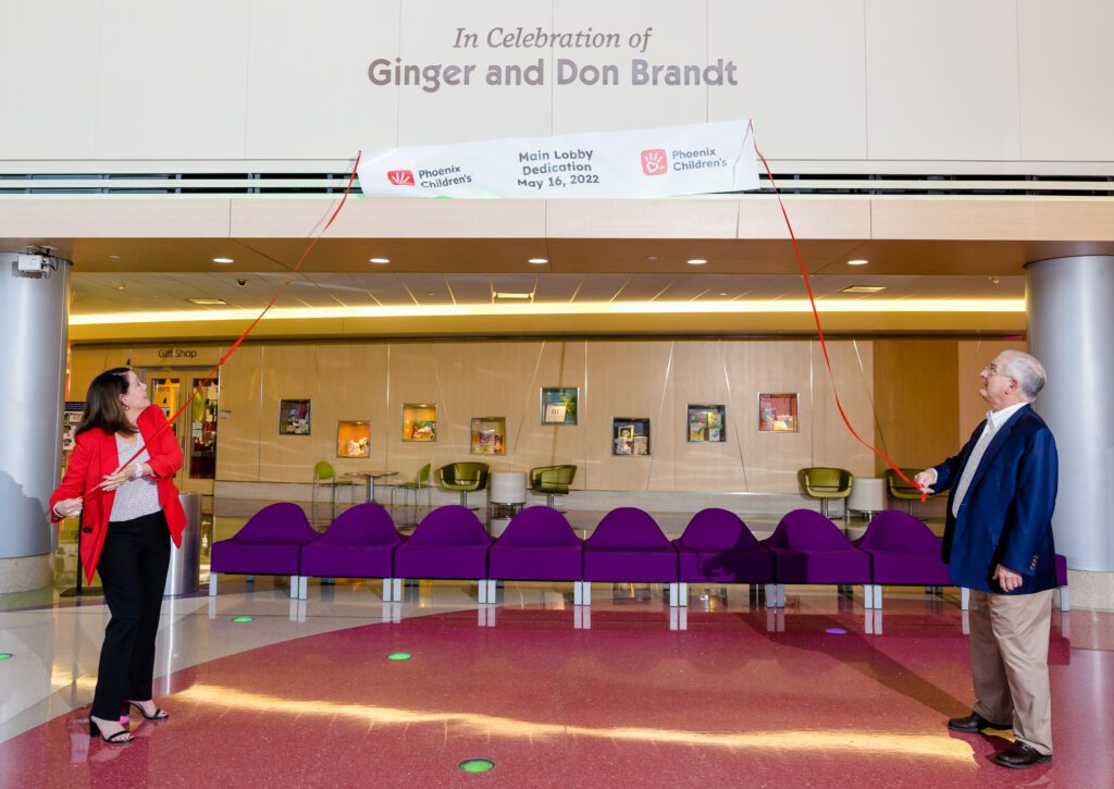 Don and Ginger pull a temporary sign off of the lobby wall at Phoenix Children's. Behind the sign there is permanent text on the hospital wall that reads, "In Celebration of Ginger and Don Brandt."