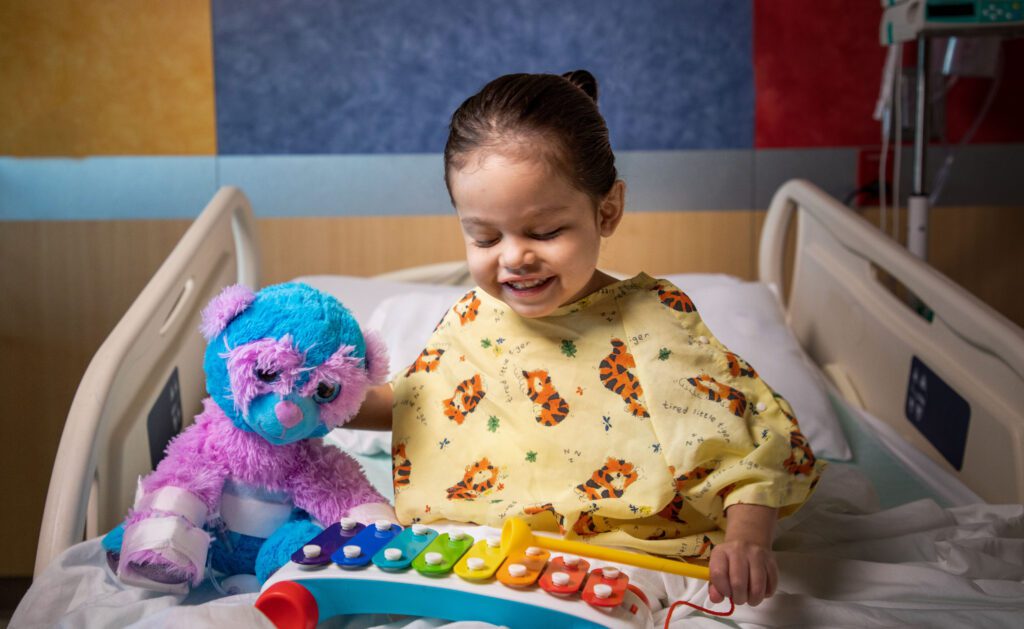 Namiko holds her teddy bear in the hospital while playing a xylophone. The teddy bear has bandages around its arm and belly.