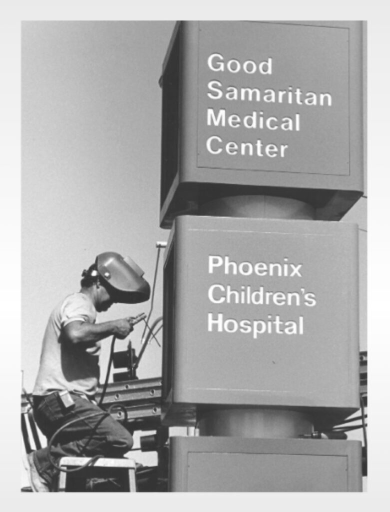 A construction worker puts up two signs, one that reads "Good Samaritan Medical Center." The other reads, "Phoenix Children's Hospital." This photo was taken in the 80s.