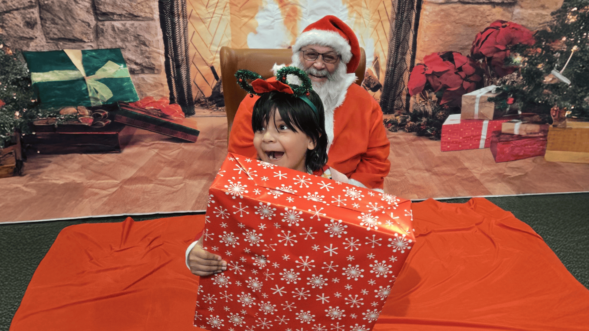 A young girl standing by Santa Claus smiles as she holds a wrapped gift.