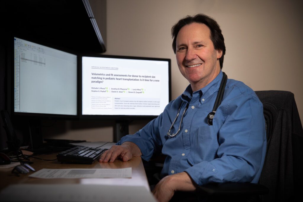 Dr. Steven Zangwill sits at his desk and poses for a photo. His research article on donor-recipient size matching in heart transplantation is up on his desktop computer screen behind him. 