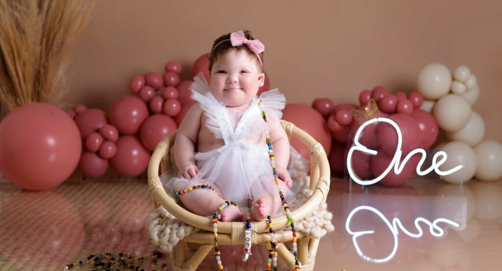 Joy on her first birthday. There are balloons in the background and she wears her beads of courage around her body