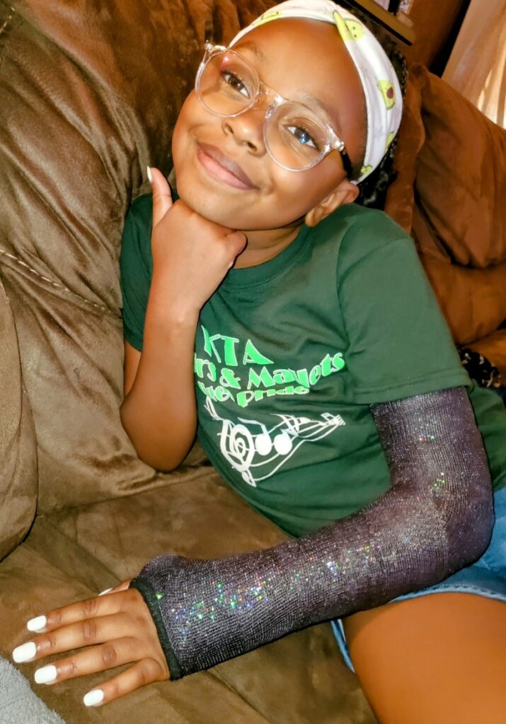 Madison poses for a photo on her couch at home. She has a cast, it's black with sparkles on it. 