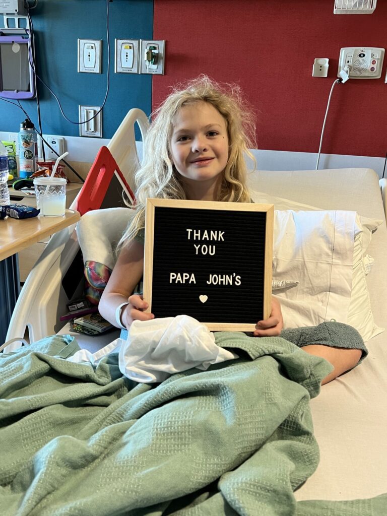 A young hospital patient holds a sign that says "Thank You, Pap John's" as she rests on her hospital bed at Phoenix Children's