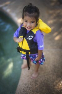 A child wearing a yellow life jacket stands next to a pool. 
