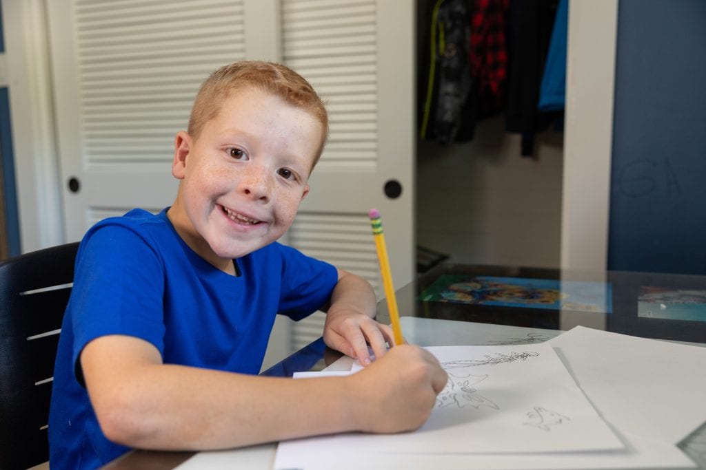 A young boy smiles as he draws a picture with pencil and paper.
