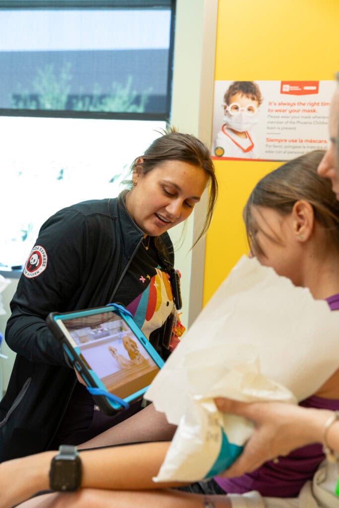 A young girl watches a video on a tablet as she sits on her mom's lap in a doctor's office. 