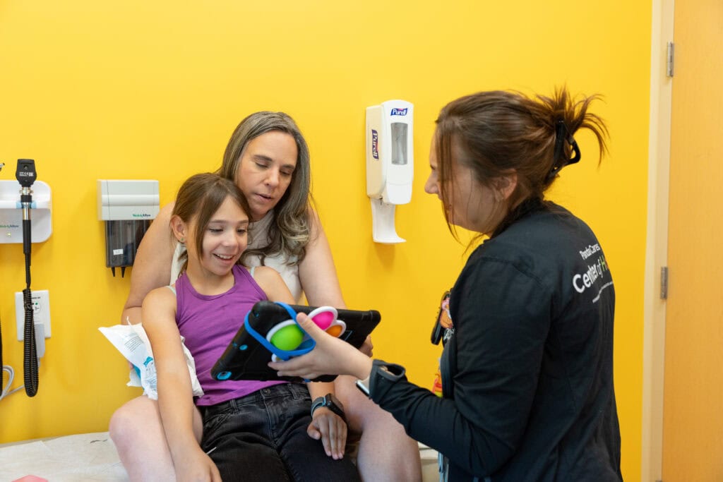 A child in a doctor's office sits on her mother's lap and looks at a tablet held by a Child Life specialist.