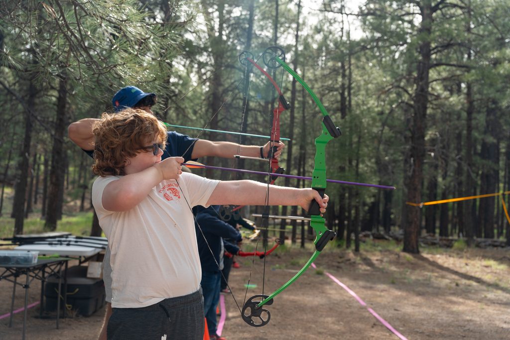 A boy in the forest stands poised to shoot with a bow and arrow.